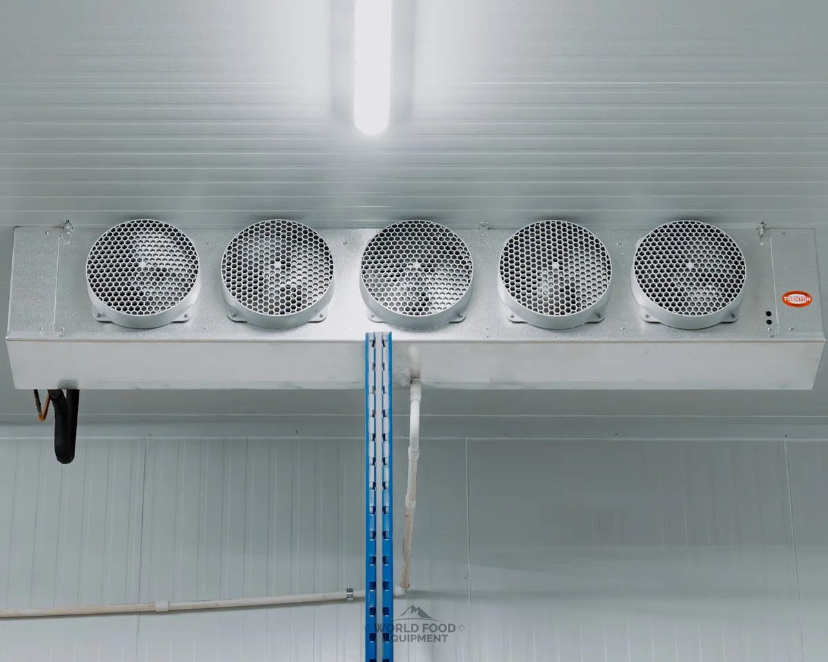 Trenton five-fan industrial evaporator ceiling-mounted on a white insulated panel wall in a finished Ontario cold storage room, blue racking upright and white drain line below, Atlas Foods install by World Food Equipment