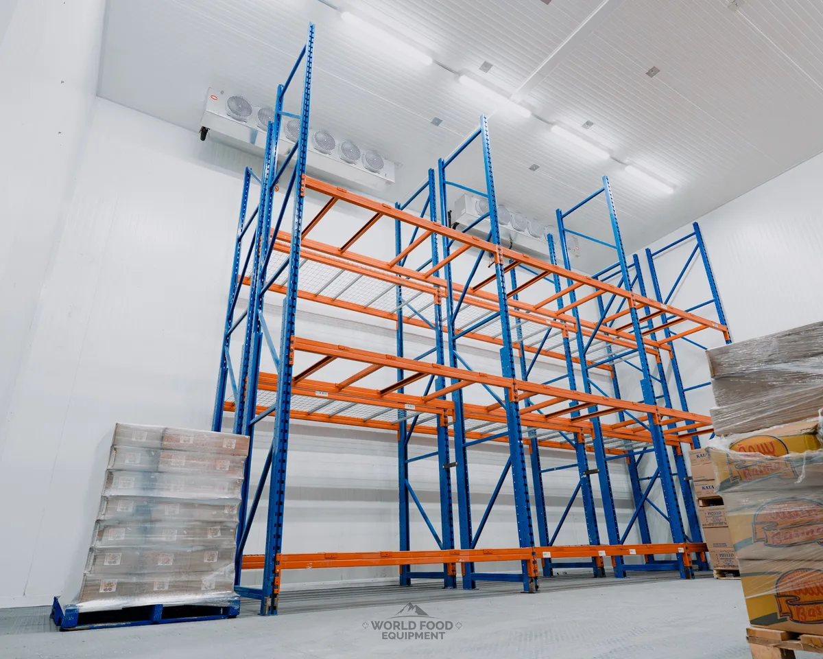 Tall blue and orange pallet racking inside a finished commercial cold storage warehouse in Ontario, two ceiling-mounted multi-fan evaporators, white insulated panel walls and ceiling, palletized product on the floor, Atlas install by World Food Equipment