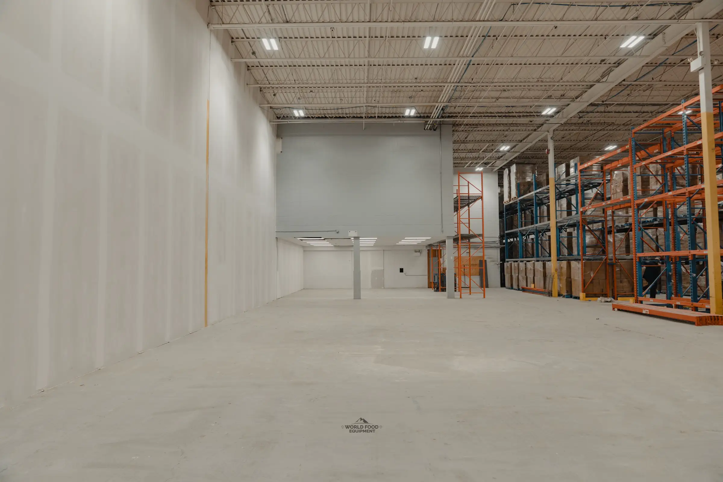 Empty Toronto warehouse bay at Atlas Foods before World Food Equipment built the cold storage: high open-web steel roof deck, bare concrete floor, drywall wall on the left and existing pallet racking on the right