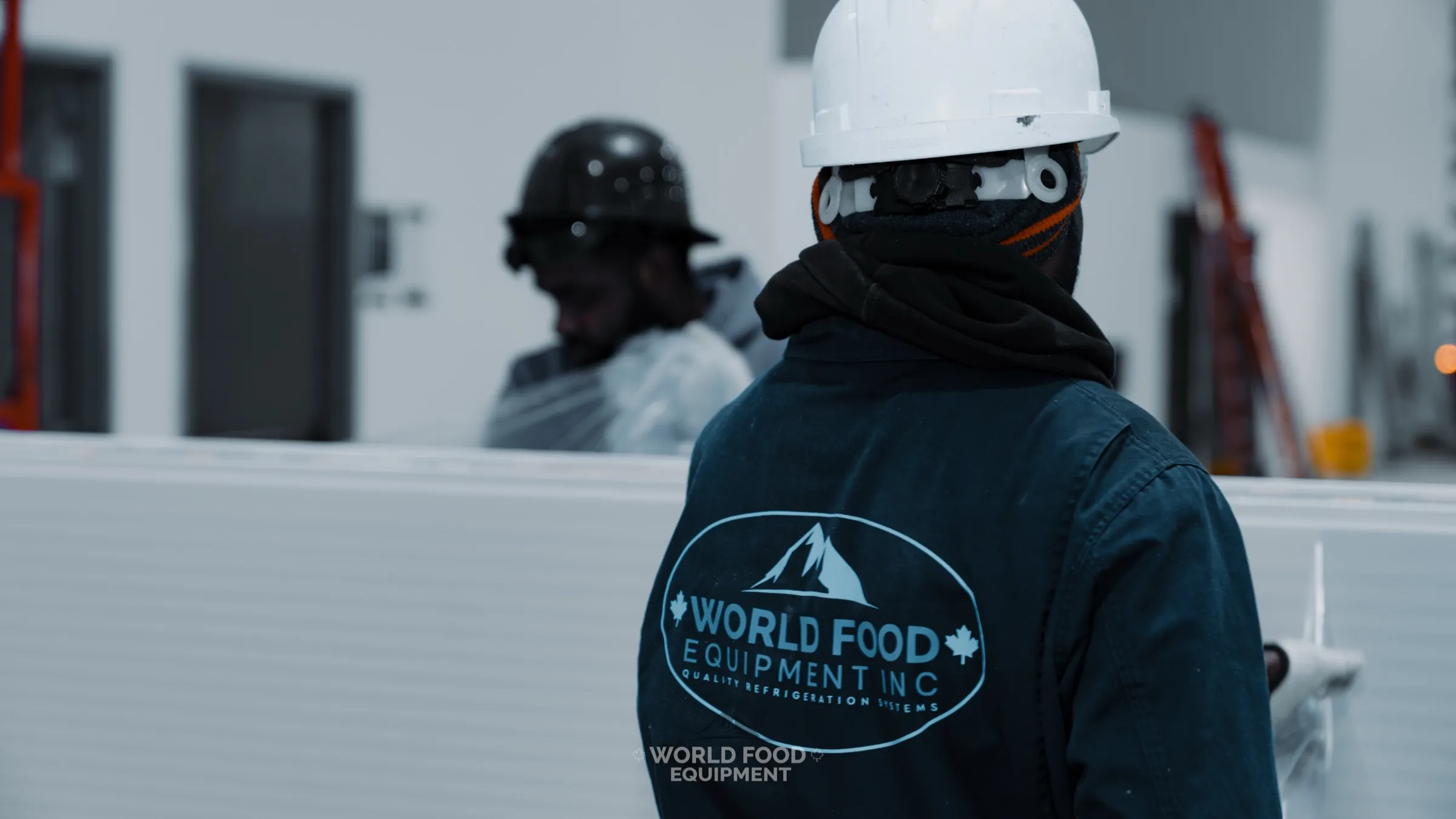 World Food Equipment crew member in a navy WFE-branded coverall and white hard hat handling a large white insulated cold storage panel on an install site, a second hard-hatted crew member working in the background, in-house WFE install crew