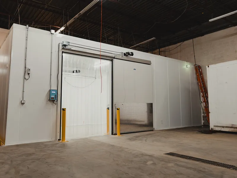 White commercial walk-in cooler exterior with a sliding insulated door, twin yellow steel bollards, a digital temperature control panel, polished concrete floor, SKOR Crestlawn install, Ontario, World Food Equipment