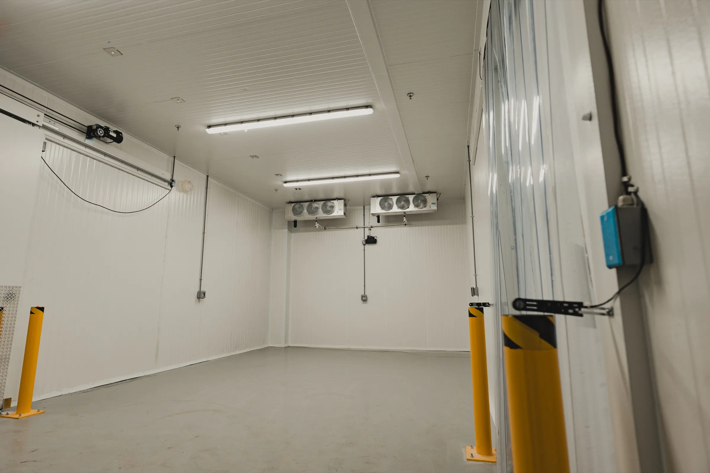 Empty interior of the 3,000 sq ft walk-in freezer at SKOR Crestlawn with dual ceiling-mounted evaporators and yellow safety bollards staged at the doorway, school food supplier freezer + processing build.