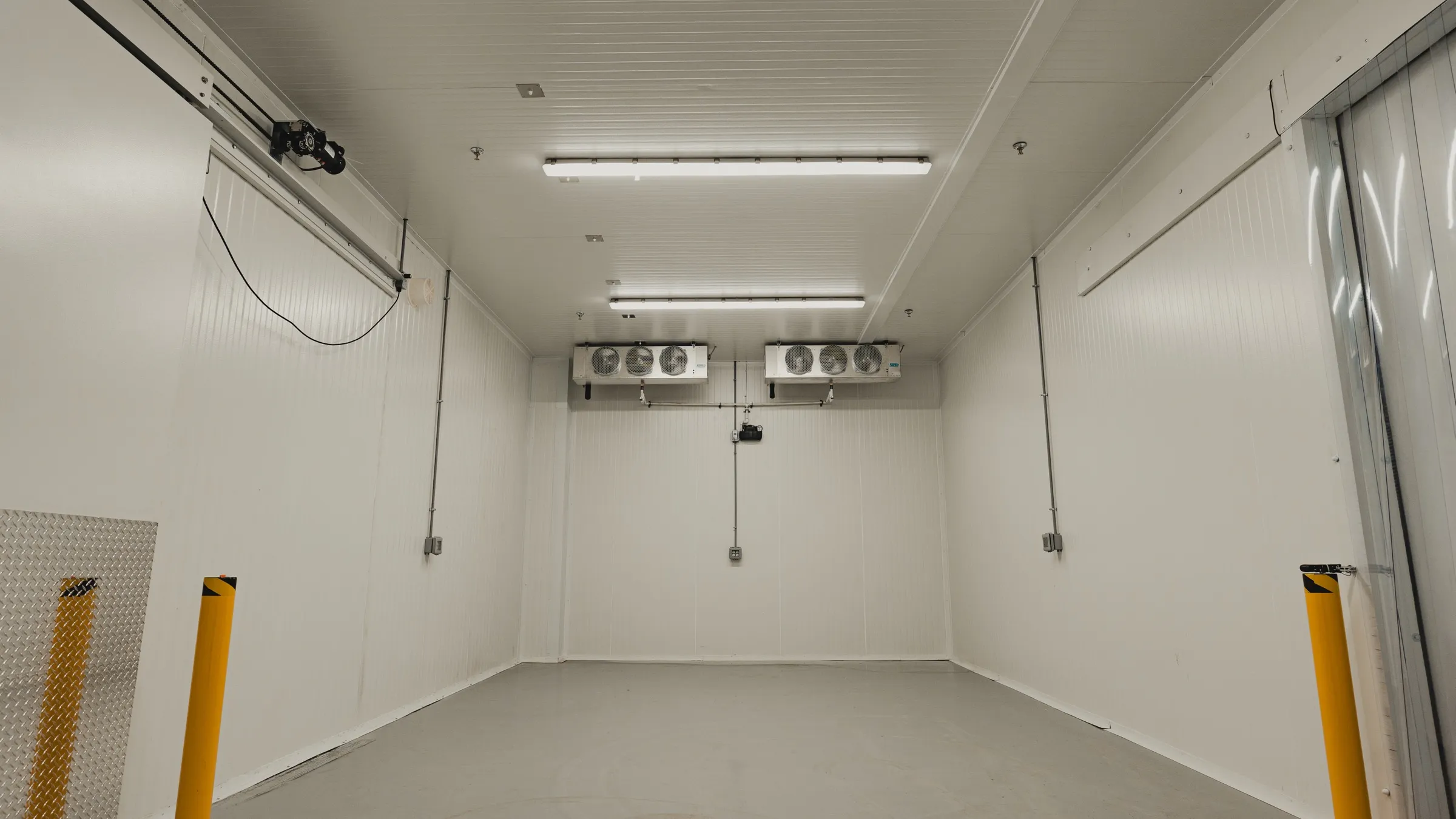 Finished commercial walk-in cold room in Ontario with white insulated panel walls and ceiling, twin three-fan ceiling evaporators on the back wall, a motorized sliding insulated door on the left, yellow safety bollards and diamond-plate kickplate at the entry, SKOR Crestlawn install by World Food Equipment