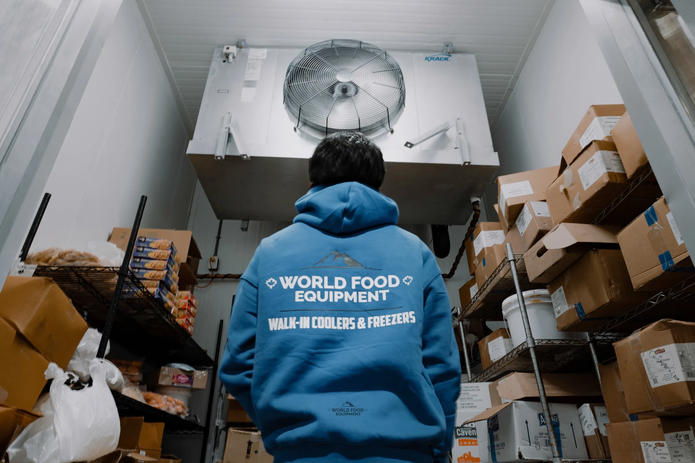 World Food Equipment crew member in a blue WFE hoodie looking up at a KRACK single-fan evaporator inside a stocked walk-in cooler at the Food Junction install in Mississauga, Ontario