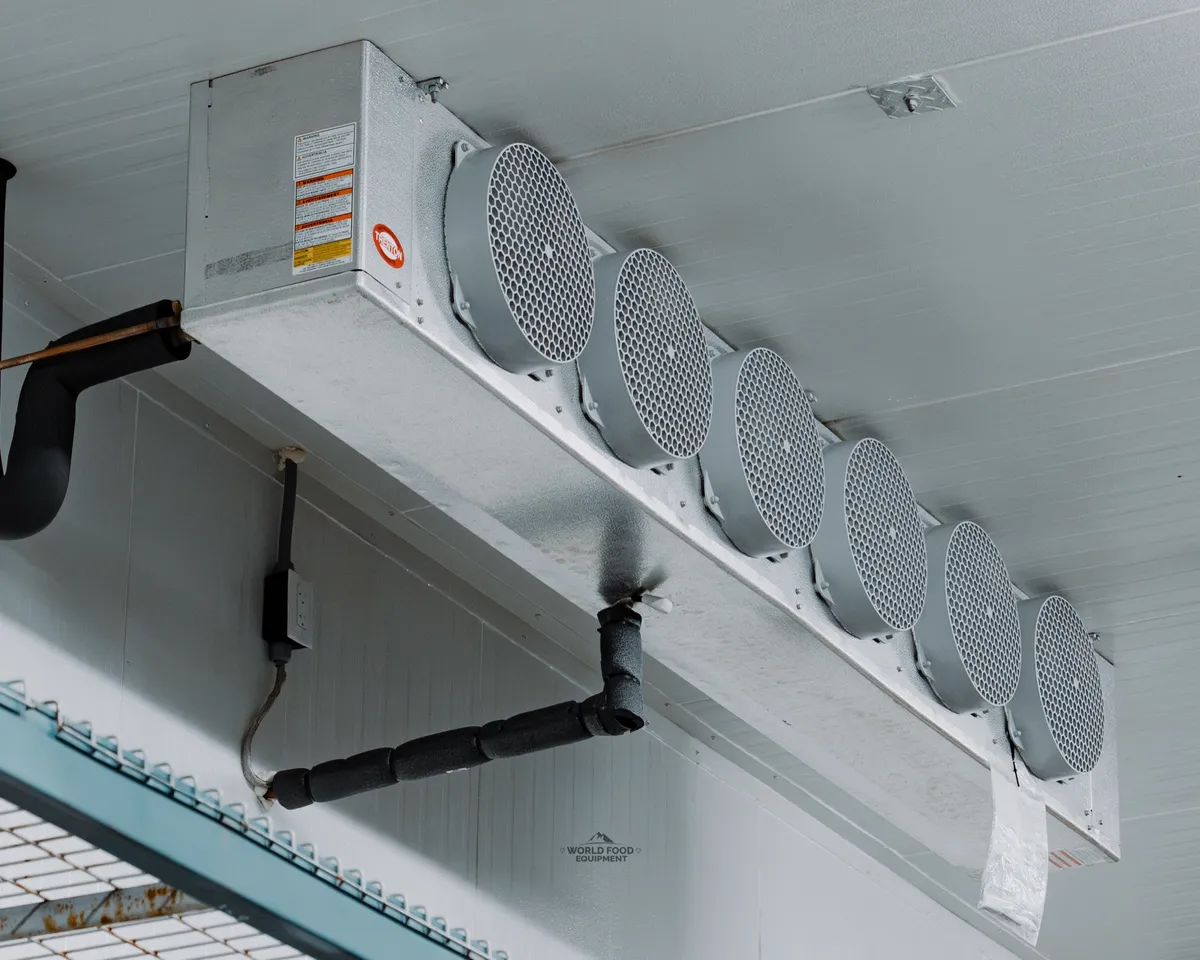 Empty finished commercial walk-in freezer interior in Ontario with two ceiling-mounted three-fan evaporators, a sliding insulated door on a top track with yellow bollards, and white insulated panel walls under LED strip lights, Food Junction install in Mississauga by World Food Equipment