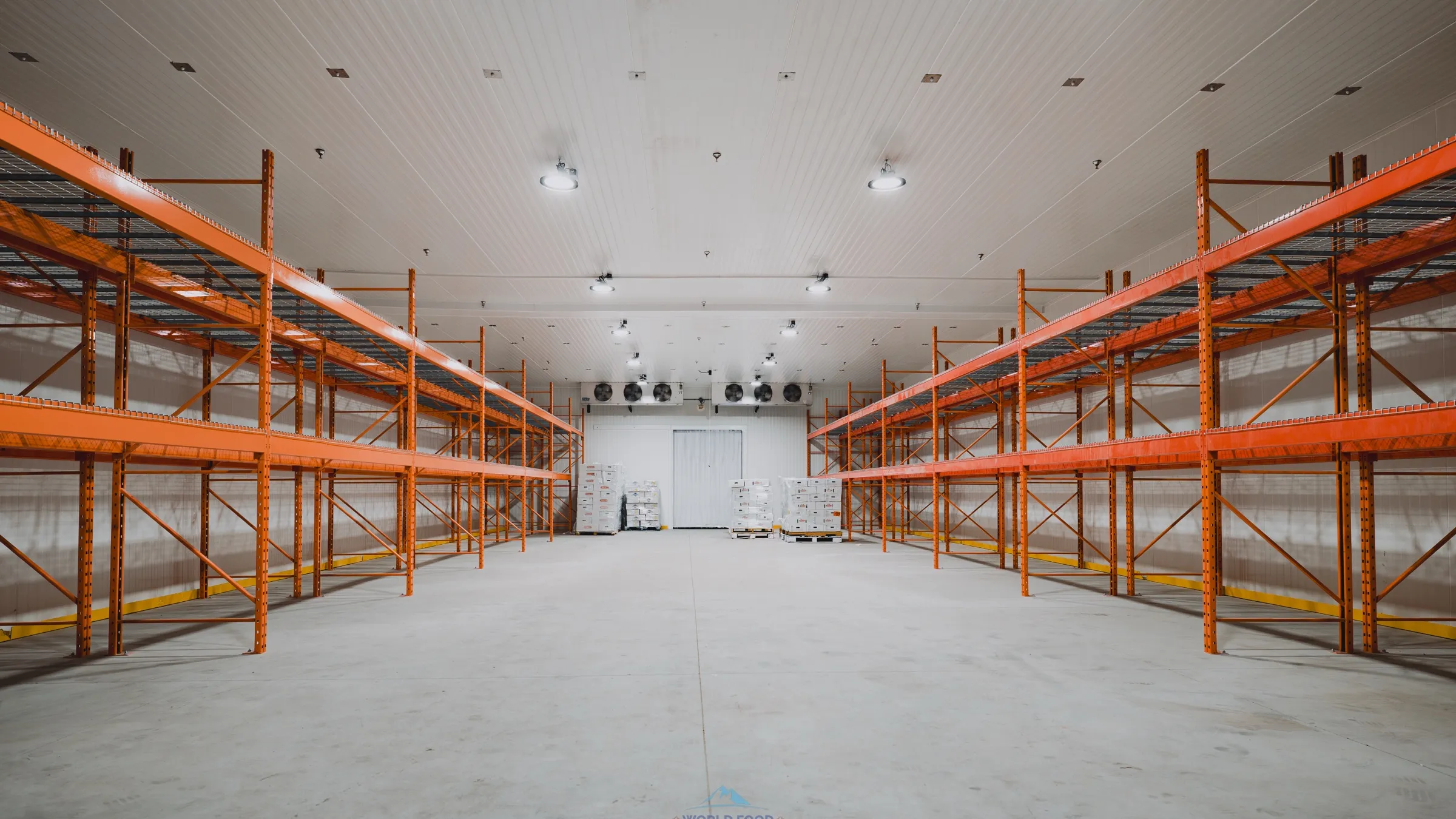Large commercial walk-in freezer warehouse in Ontario with orange pallet racking down both sides, two ceiling-mounted multi-fan evaporators over the back door, palletized frozen product staged on the floor, white insulated panel walls and ceiling, halal meat processor install by World Food Equipment