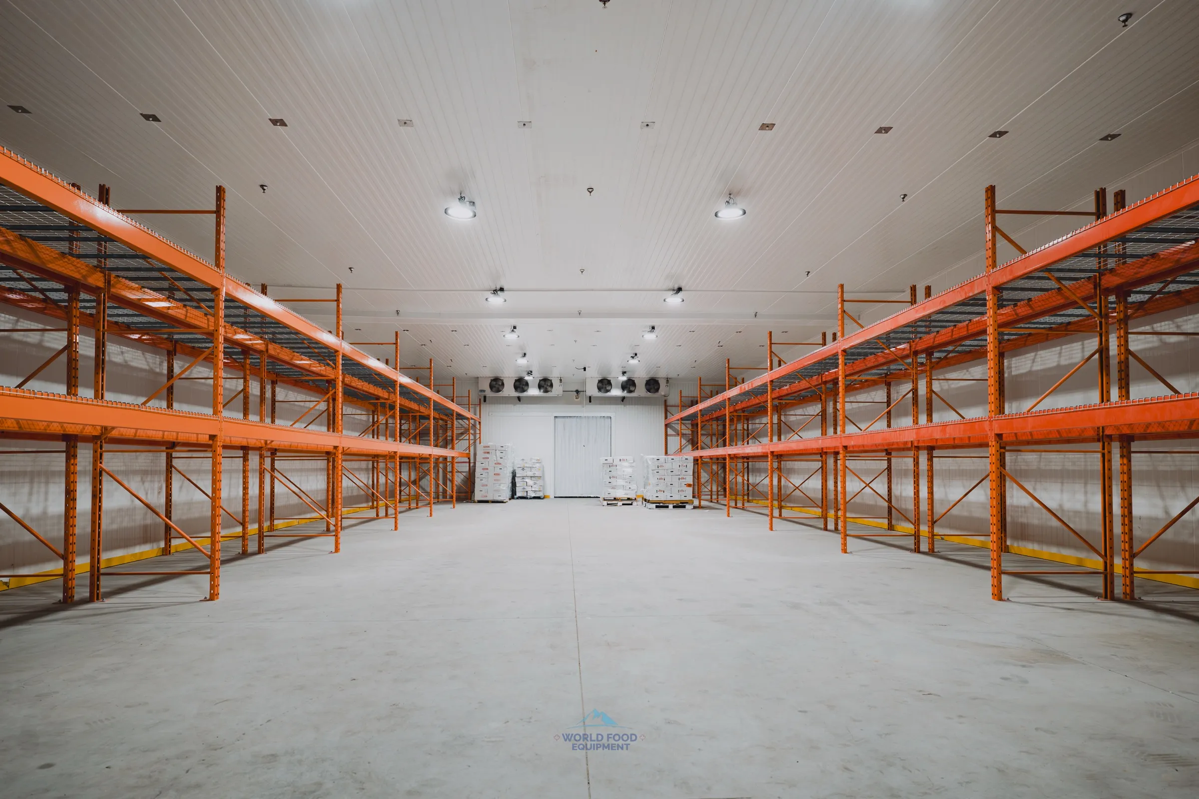 Wide industrial cold storage room in a halal meat processing facility, built by World Food Equipment in Ontario. Twin runs of orange industrial pallet racking on both sides of a central forklift aisle, six ceiling-mounted KeepRite evaporator units lined across the back wall, insulated panel walls and ceiling, polished concrete floor