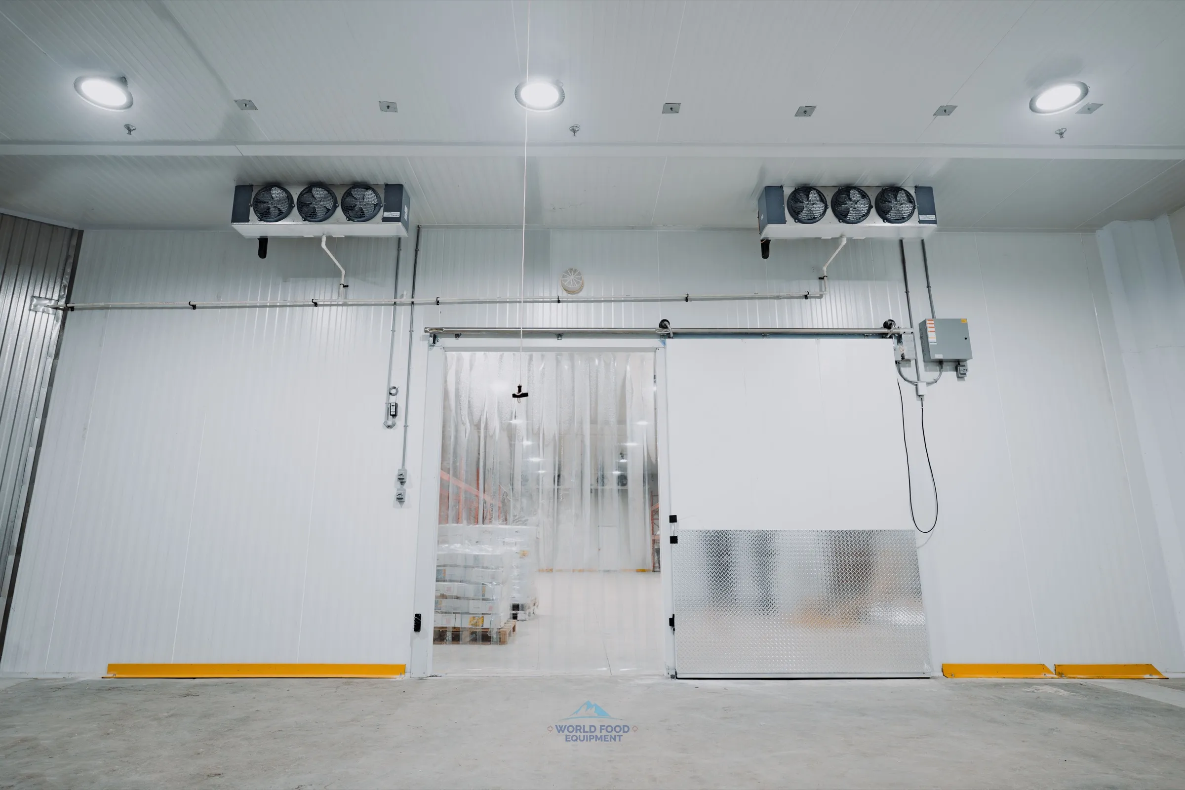 Operational commercial walk-in freezer at an Ontario meat-processing facility with dual ceiling-mounted low-temp evaporators, sliding insulated door open with vinyl strip curtain pulled aside, stacked product pallets inside, World Food Equipment install