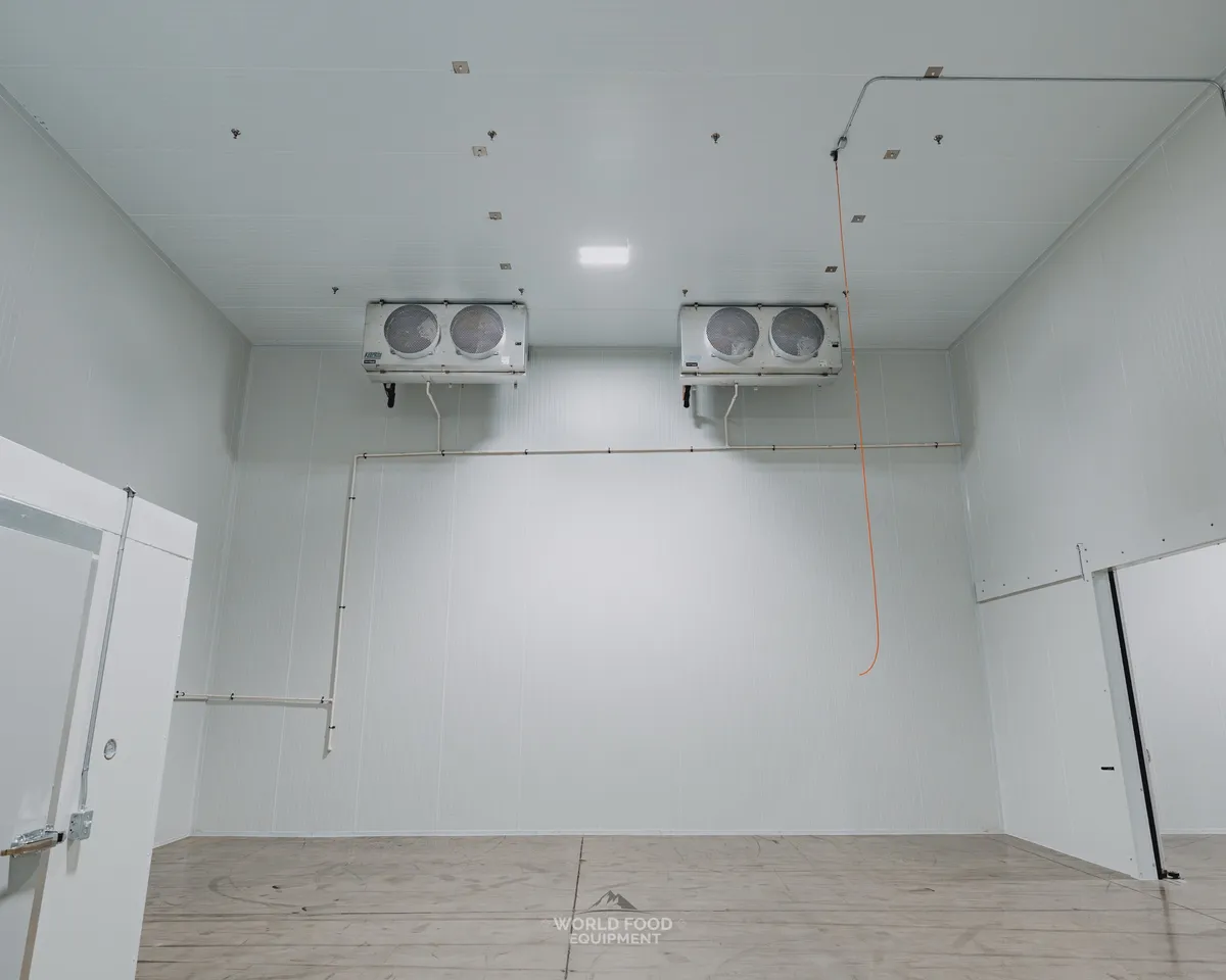 Finished commercial cold storage room in Mississauga with white insulated panel walls, an insulated swing door with a temperature gauge and World Food Equipment decal, a wide sliding insulated door opening, and twin-fan ceiling evaporators, Ontario walk-in installation by World Food Equipment
