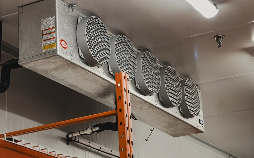 Trenton five-fan evaporator ceiling-mounted above orange pallet racking in a finished Ontario cold room, with the white PVC condensate drain line and insulated refrigerant lines routed along the panel wall, Stainless Steel project by World Food Equipment