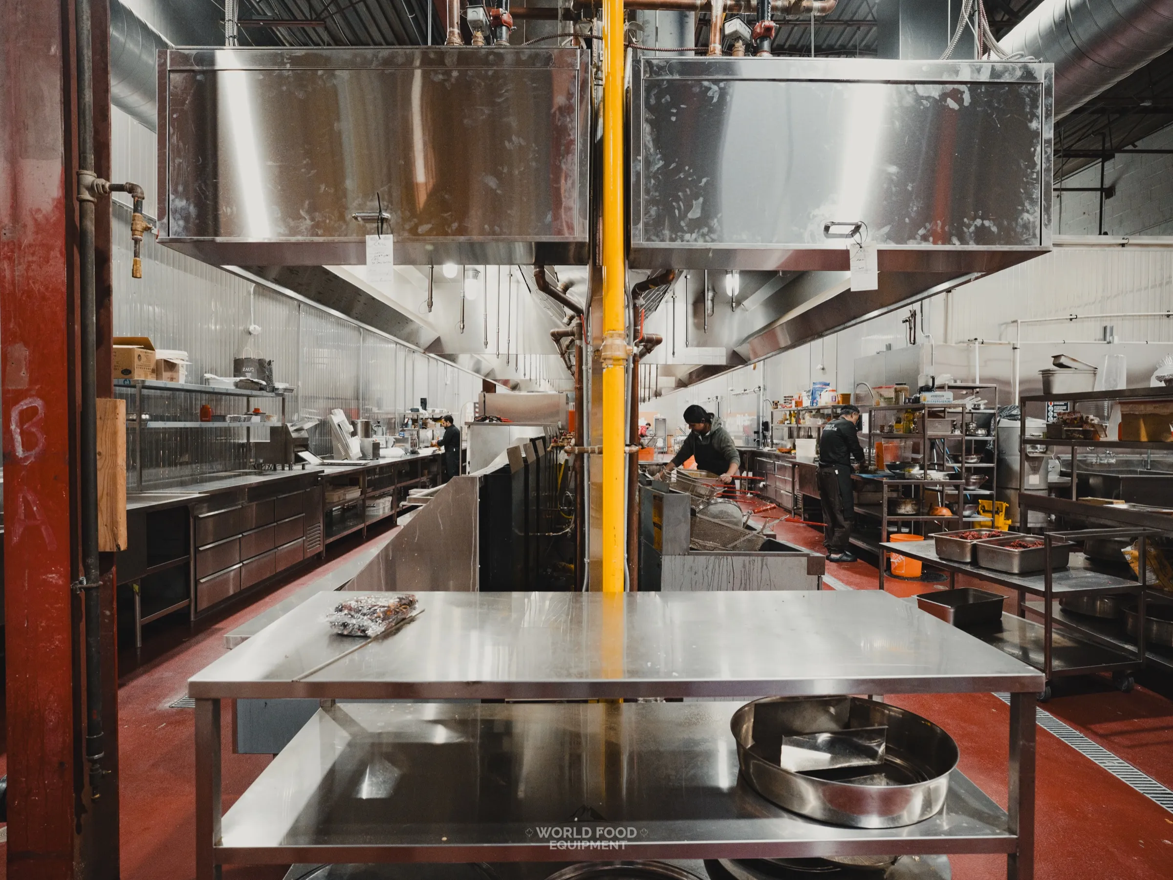 Commercial kitchen at the Stainless Steel project in Ontario with stainless exhaust hoods over the cooking line and a yellow-painted gas line dropping to the equipment, World Food Equipment