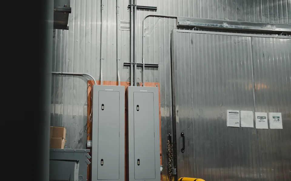 Two grey electrical distribution panels mounted on a plywood backboard beside a stainless-clad walk-in freezer door, EMT conduit runs rising to the ceiling, at the Stainless Steel project in Ontario, World Food Equipment