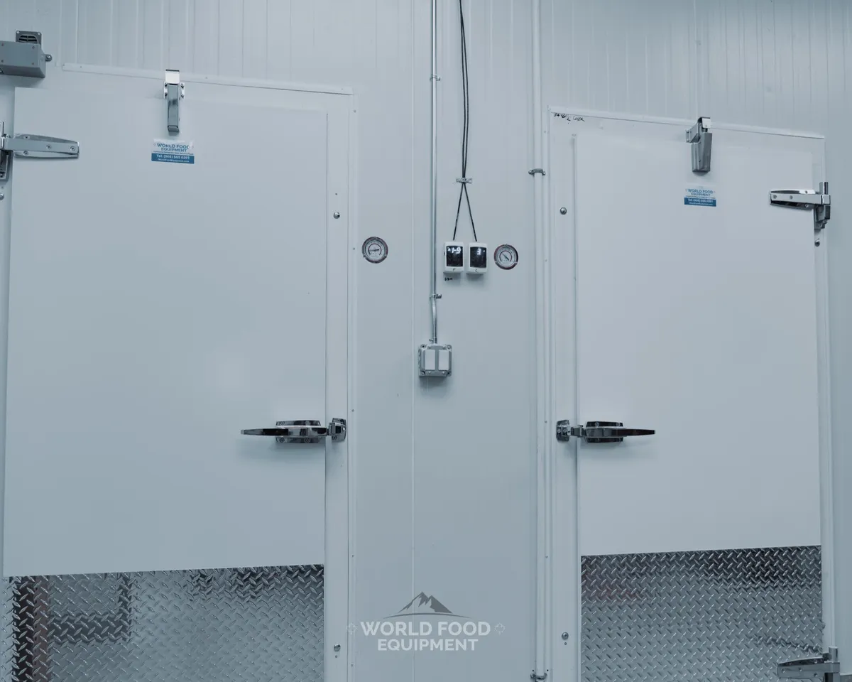 Two finished white insulated walk-in doors side by side, a cooler and a freezer, with heavy-duty hinges and latches, diamond-plate kick panels along the bottom, and a temperature gauge mounted on the panel between them, Ontario commercial walk-in cooler and freezer installation by World Food Equipment
