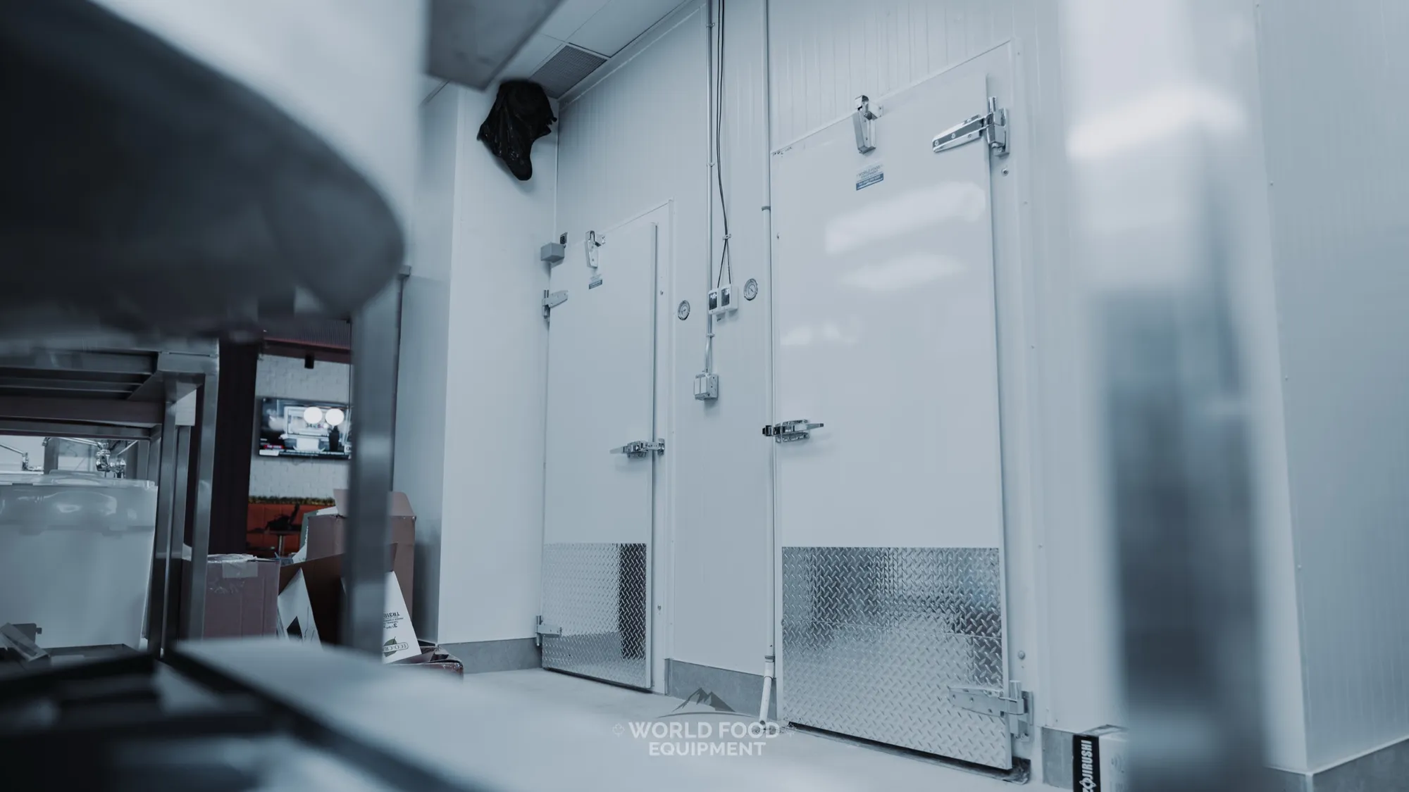 The Bagel House back-of-house in Toronto with the walk-in cooler and freezer doors side by side on one white panel wall, diamond-plate kickplates, controllers between the doors, stainless prep equipment in the foreground and the café dining room visible beyond.