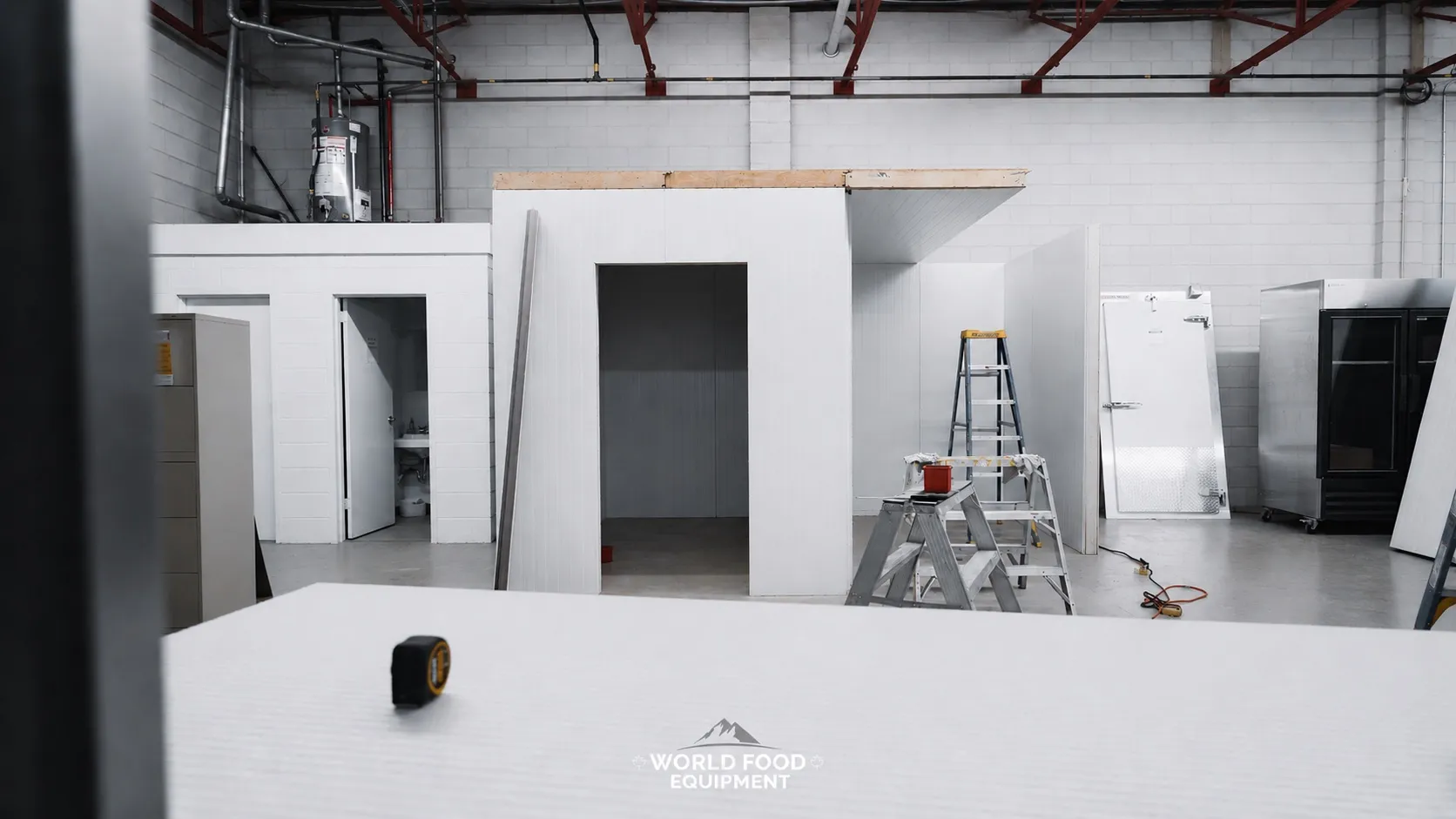 Wide interior view of a commercial warehouse with multiple finished stand-alone walk-in cooler envelopes installed on the left and center, a wide doorway opening into a finished cold storage room at the center, a tall white insulated reach-in unit and a black commercial reach-in cooler visible on the right, yellow A-frame step ladder and scissor lift staged in the room, foreground panel with a tape measure on top, concrete-block warehouse walls and exposed overhead piping, polished concrete floor, new commercial walk-in coolers and freezers built and installed by World Food Equipment, Ontario