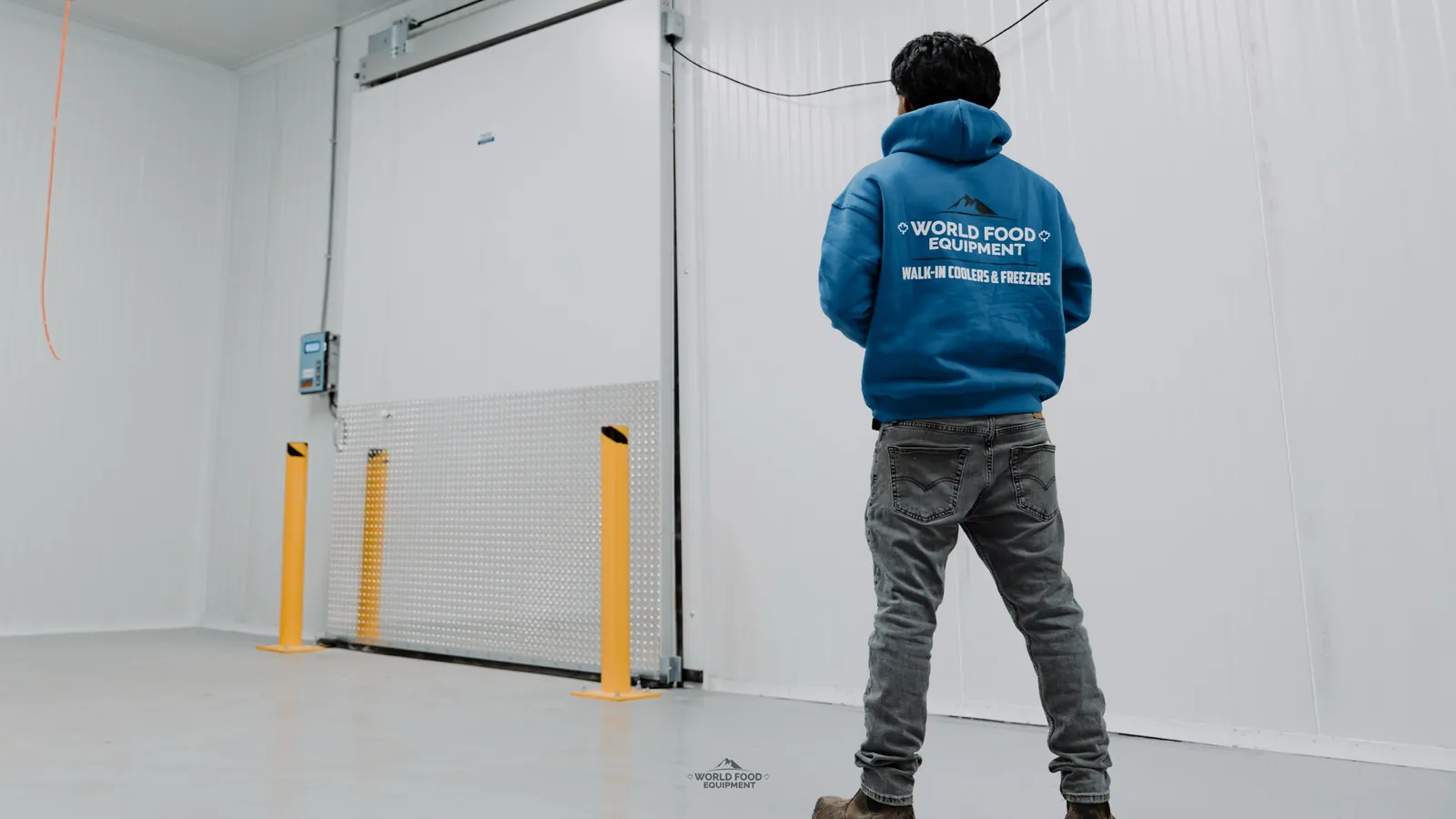 World Food Equipment crew member in front of a motorized sliding walk-in freezer door with a diamond-plate kickplate and yellow steel bollards at the SKOR Crestlawn install in Ontario