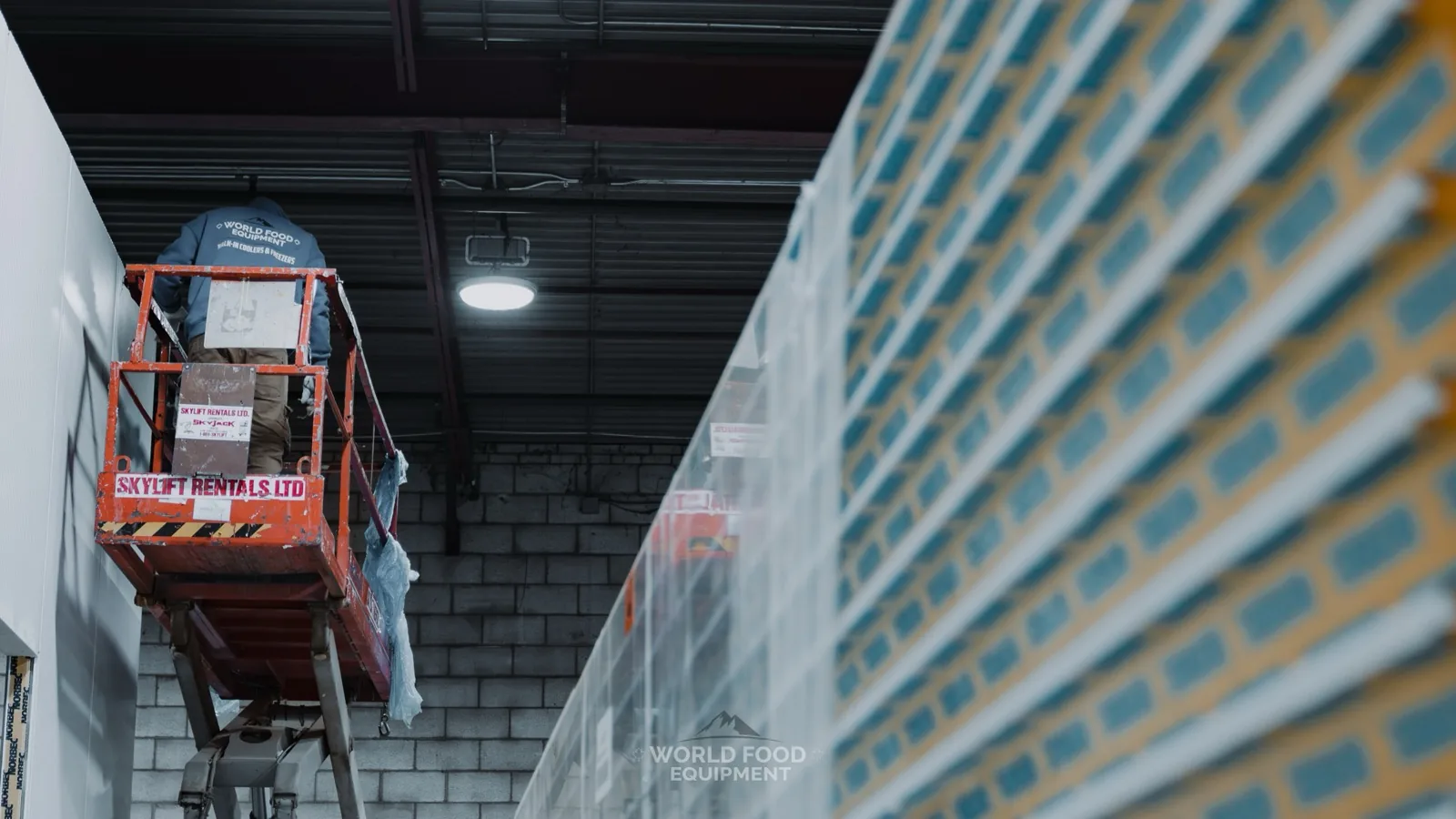 World Food Equipment installer on an orange scissor lift setting white insulated wall panels, with stacked Norbec-wrapped panels in the foreground, Ontario cold storage build