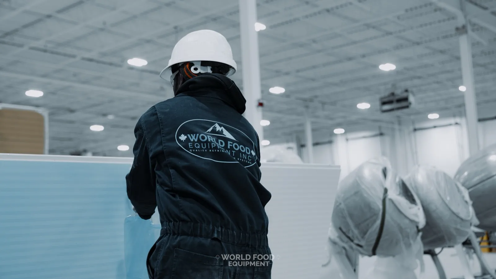 World Food Equipment crew member in a white hard hat and WFE coveralls handling a light-blue insulated walk-in cooler panel in a bright Ontario warehouse, with shrink-wrapped refrigeration equipment staged behind